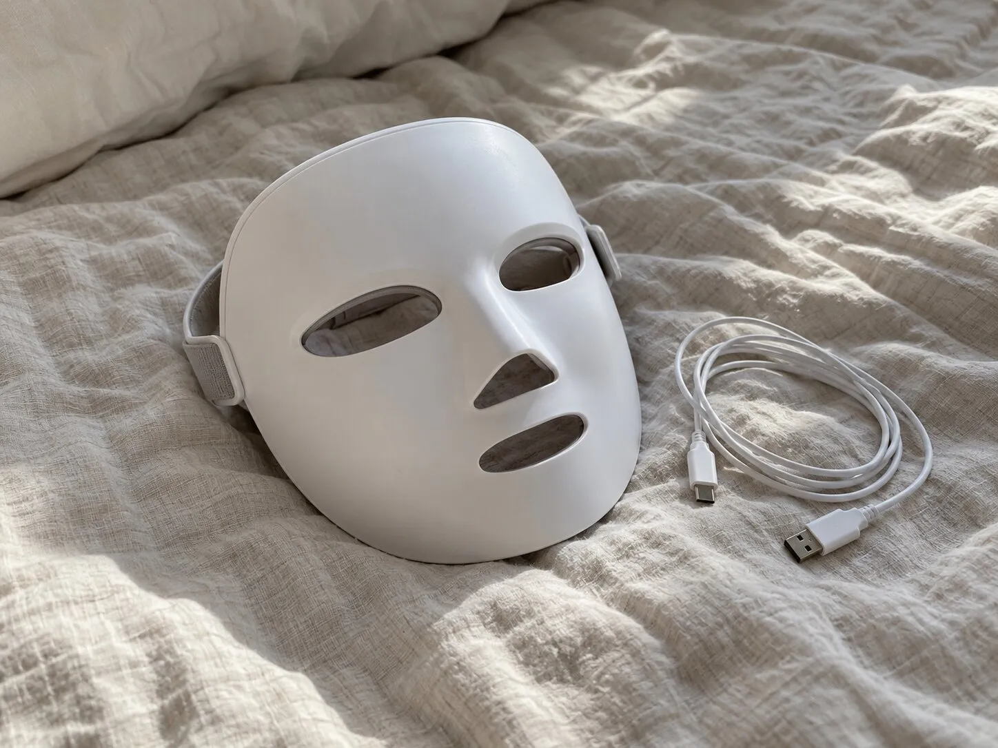 An at-home LED light therapy mask and its charging cable on a bed